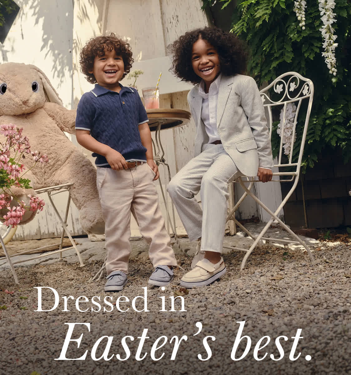 The image features two young individuals, a boy and a girl, standing in a garden setting surrounded by flowers and a large Easter bunny statue. They appear to be dressed in their Easter best, with the boy wearing a polo shirt and the girl wearing a white outfit.