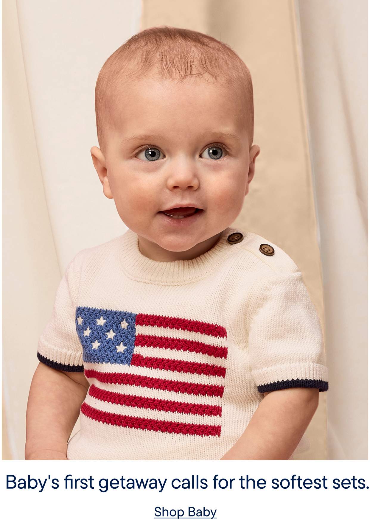 A smiling baby wearing a white sweater with an American flag design, standing in front of a light-colored wall.