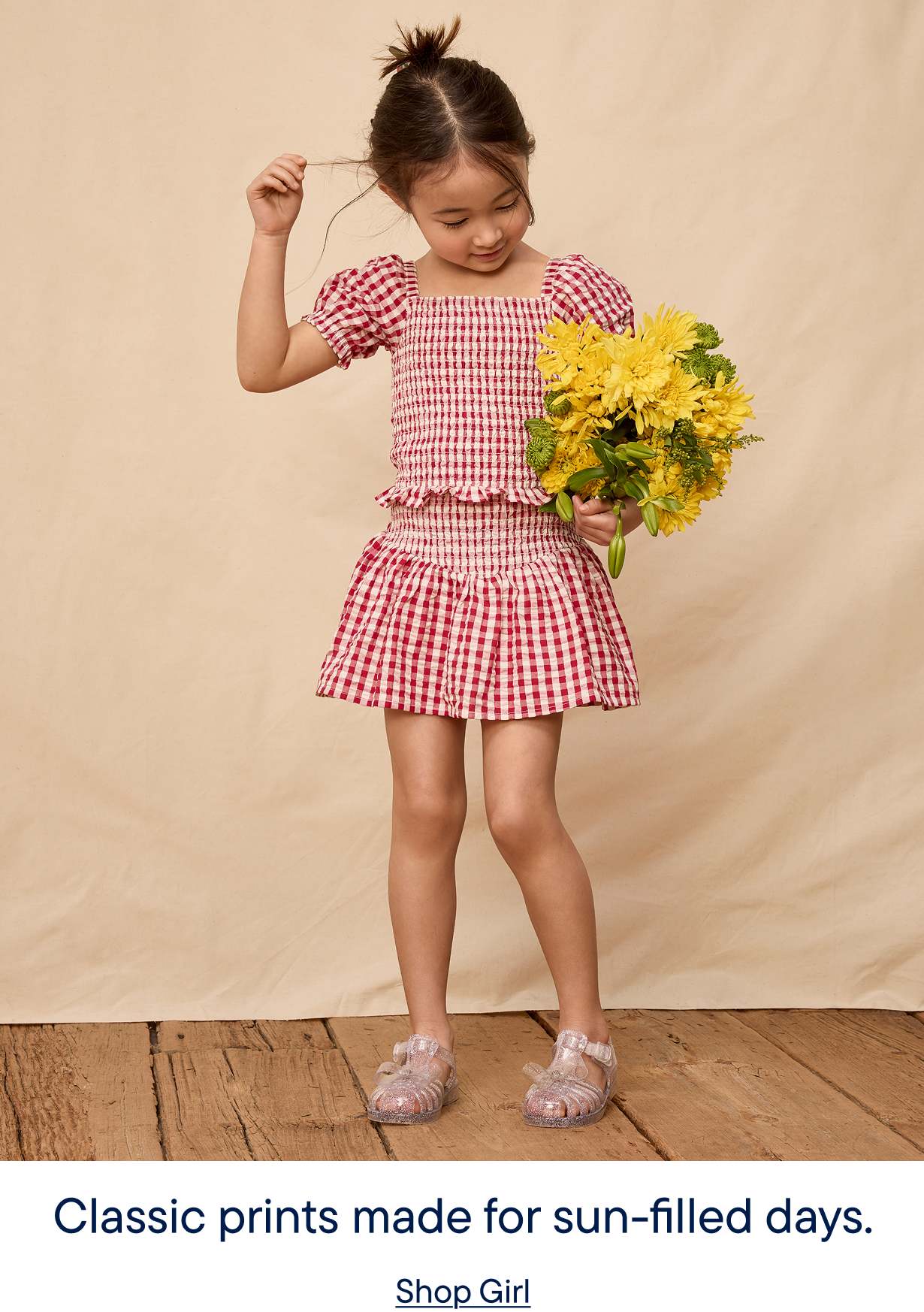 A young girl in a red and white checkered dress stands holding a bouquet of yellow flowers, with a plain beige background and wooden floor visible.