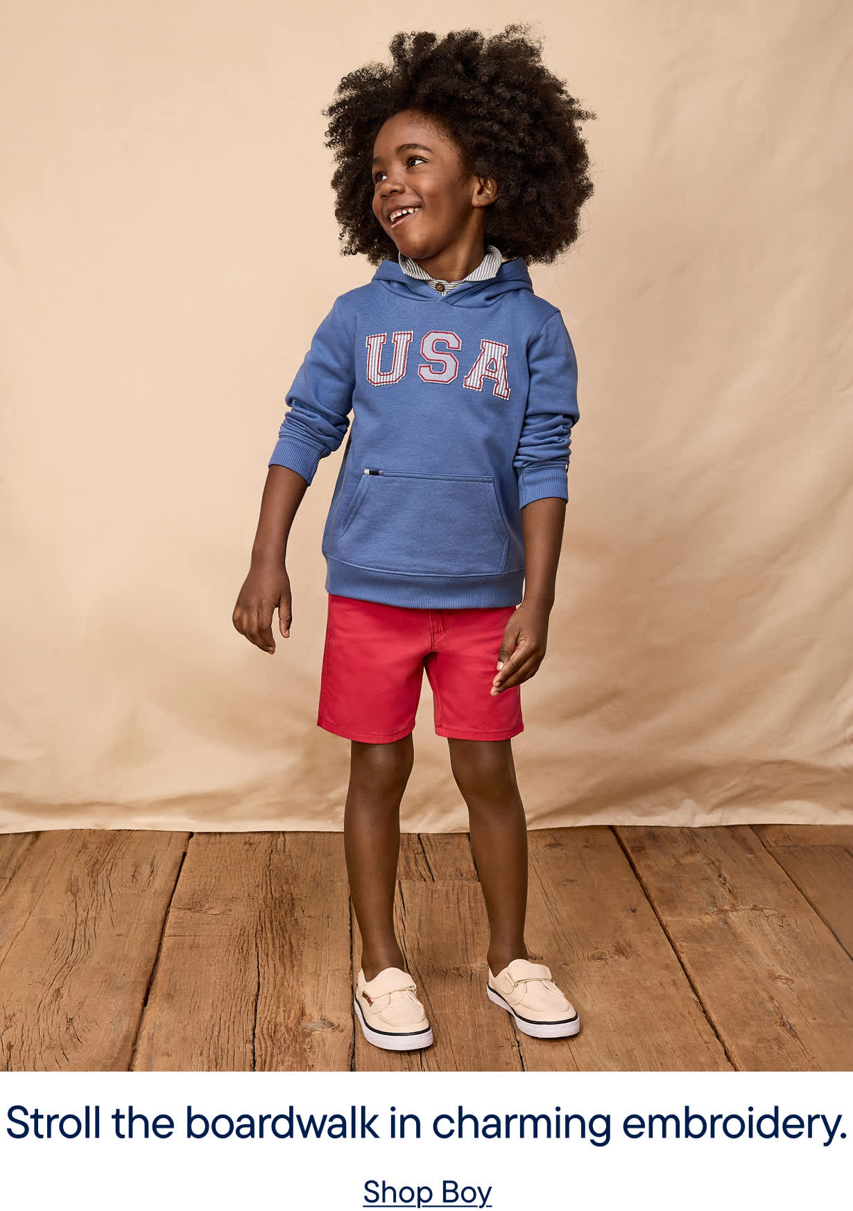 A young child with curly hair wearing a blue "USA" sweatshirt and red shorts stands on a wooden floor against a beige background.