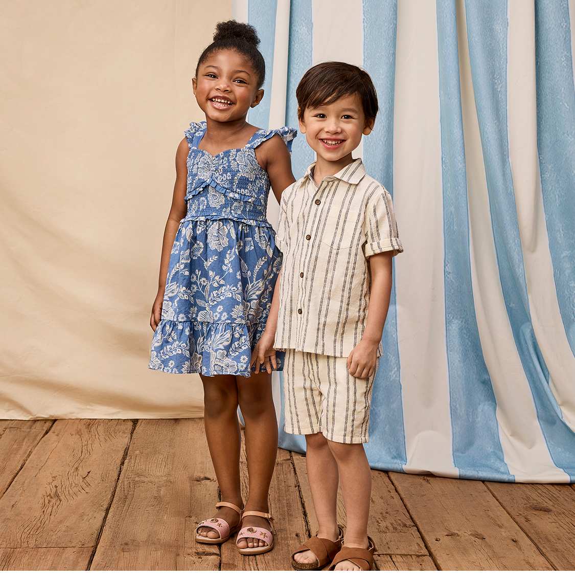Two young individuals, a girl and a boy, are standing together in front of a blue and white striped curtain backdrop. The girl is wearing a patterned dress, while the boy is wearing a striped shirt. In the background, there is a sign advertising "Iconic Summer Looks" with a promotional offer.
