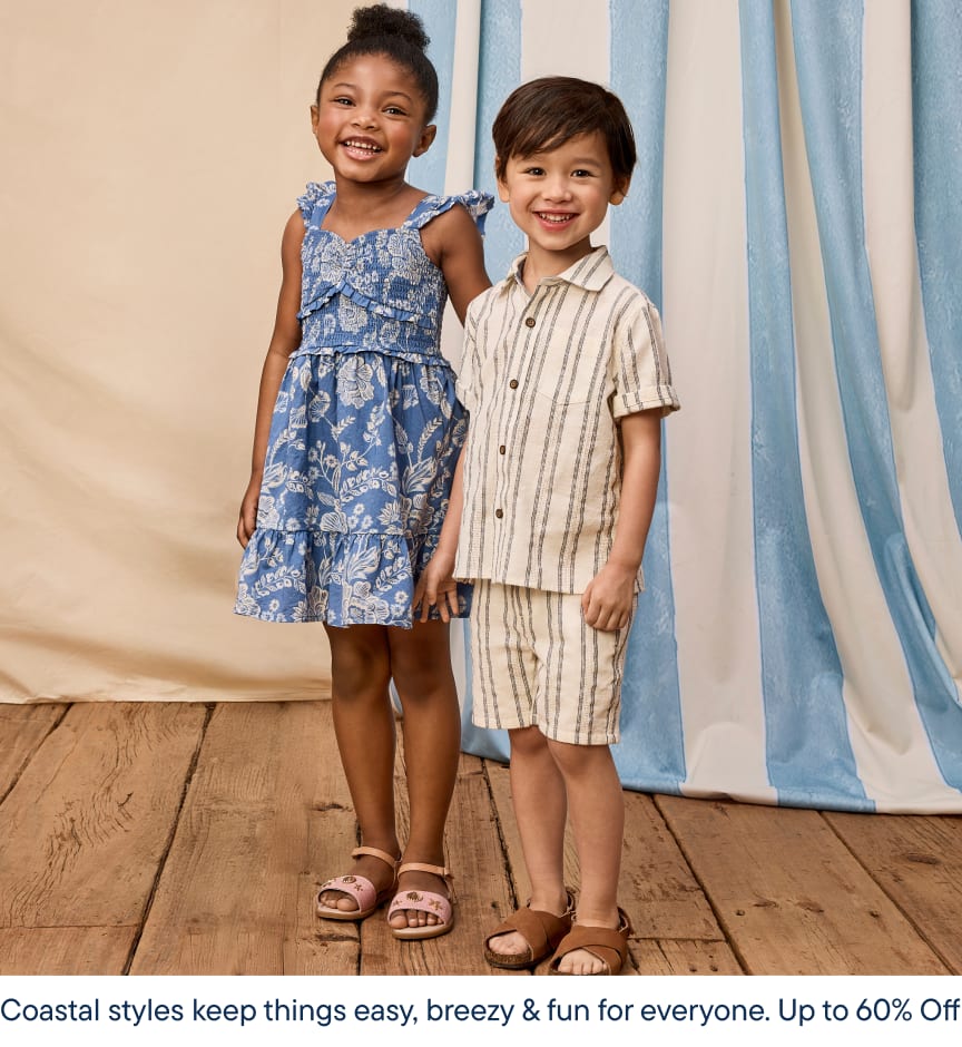 Two young children, a girl in a blue floral dress and a boy in a striped shirt, are standing together on a wooden floor with blue curtains in the background. The image has a coastal, breezy feel, suggesting a fun and easy-going style.