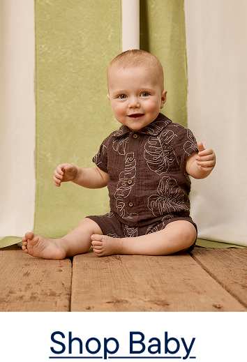 A smiling baby in a brown leaf-print outfit sits on a wooden floor in front of a green and white curtain.