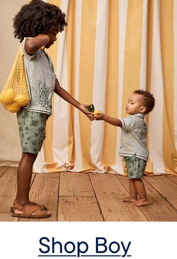 Two young boys in matching outfits stand on a wooden floor, with the older boy handing a lemon to the younger, against a backdrop of yellow and white striped curtains.