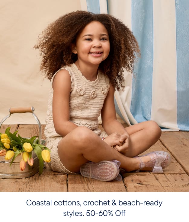 A smiling young girl in a crochet outfit sits on a wooden floor with a bucket of tulips to her left, against a backdrop of blue and white striped curtains.