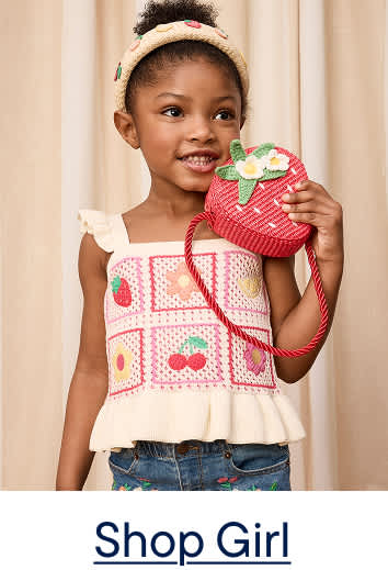 A young girl with a strawberry purse smiles in front of a cream-colored, pleated backdrop.