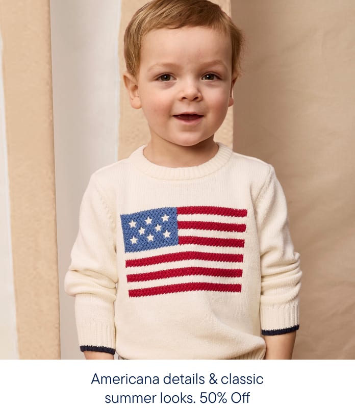 A smiling toddler in a cream sweater with an American flag design stands against a textured beige background.