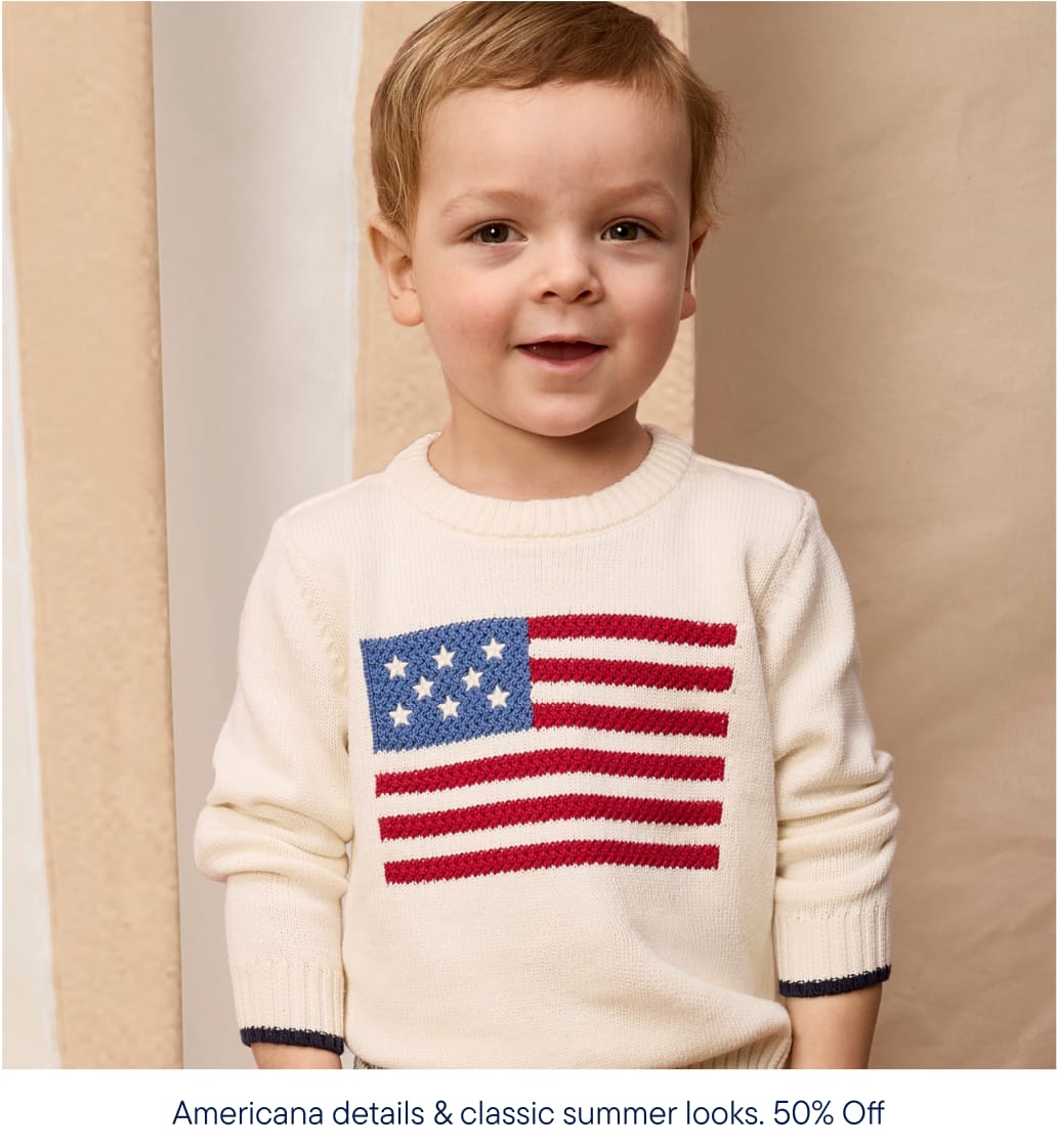 Un niño pequeño sonriente, con un suéter color crema con el diseño de la bandera estadounidense, posa sobre un fondo neutro y texturizado.