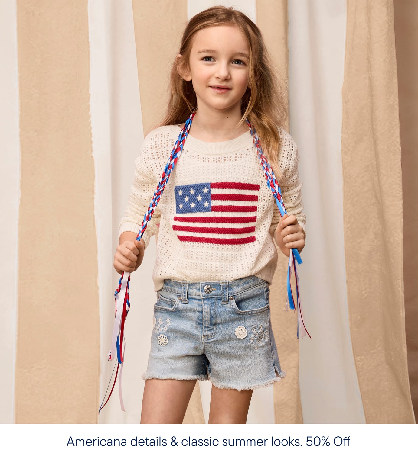 En primer plano, una joven con el pelo trenzado luce un suéter color crema con el diseño de la bandera estadounidense y pantalones cortos vaqueros, sobre un fondo de cortinas a rayas beige y blancas.