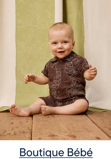 Un bébé souriant, vêtu d'une tenue marron à motifs de feuilles, est assis sur un plancher en bois devant un rideau vert et blanc.