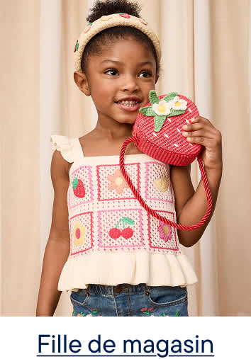 Une jeune fille, un sac à main en forme de fraise à la main, sourit devant un fond plissé couleur crème.