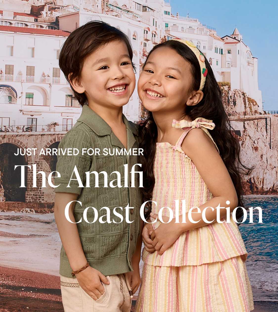 Two smiling children in summer clothing stand in the foreground with a picturesque Italian coastal town in the background.