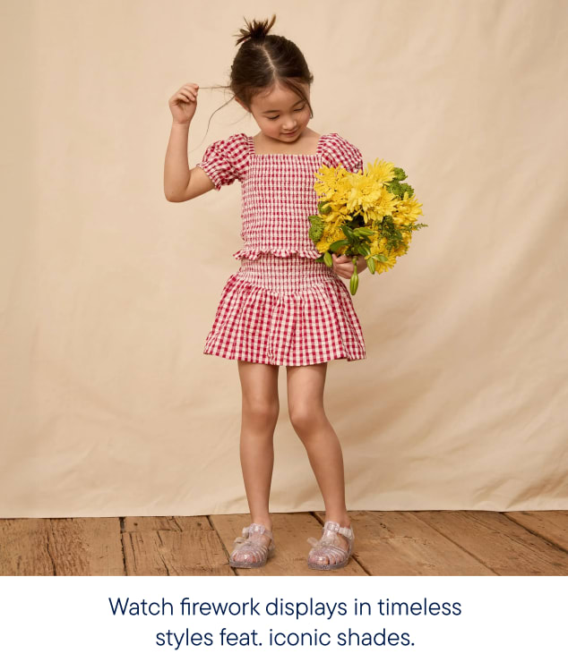 A young girl in a red gingham outfit holds a bouquet of yellow flowers against a plain backdrop.