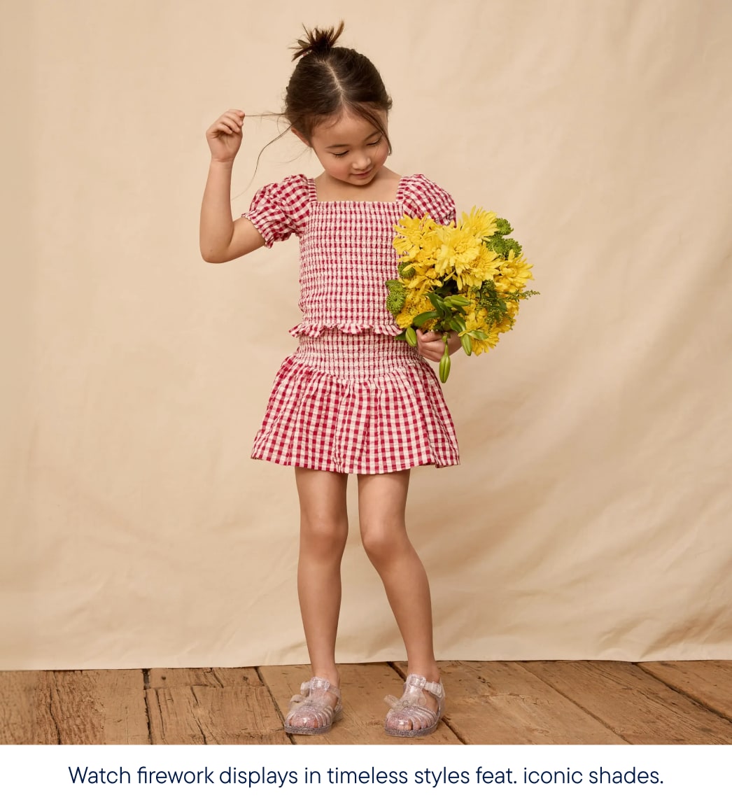 A young girl in a red gingham outfit holds a bouquet of yellow flowers against a plain backdrop.