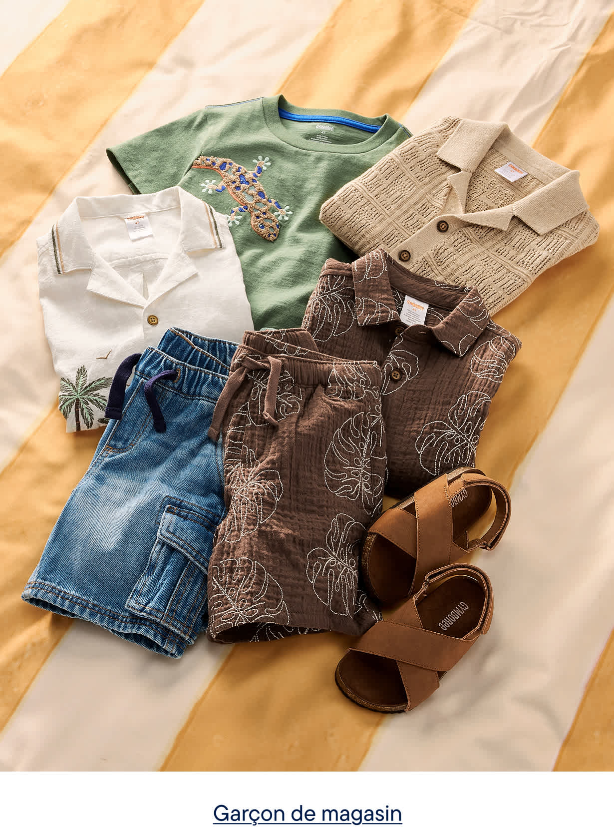 Une collection de vêtements d'été pour garçons, comprenant des shorts, des chemises et des sandales, est disposée sur une surface rayée jaune et blanche.