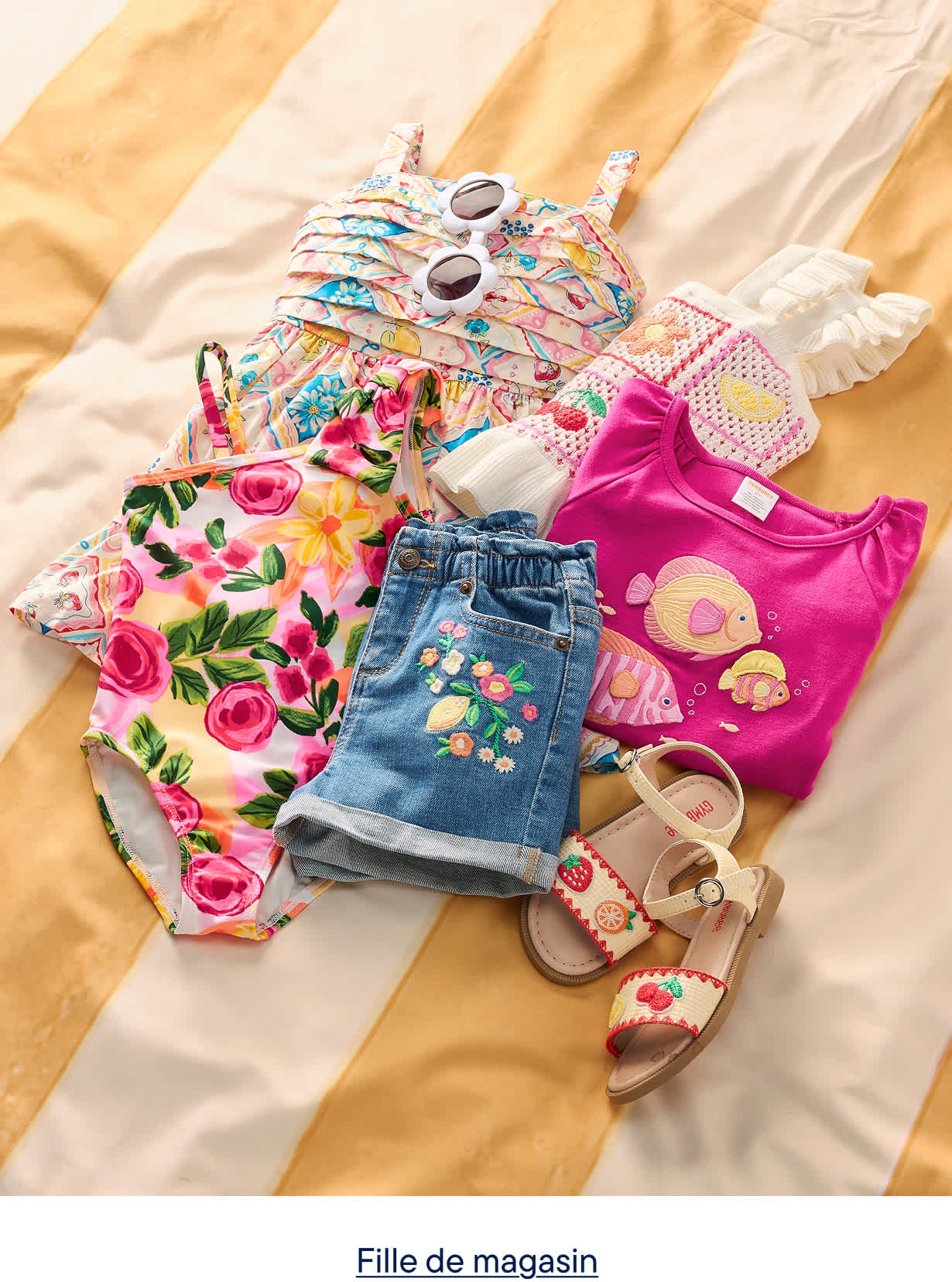 Une collection de vêtements d'été colorés pour enfants, comprenant un maillot de bain à fleurs, un short en jean, un t-shirt rose et des sandales, est disposée sur une couverture à rayures jaunes et blanches.