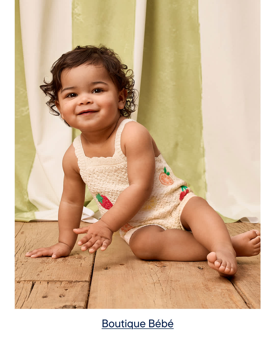 Un bébé souriant, vêtu d'une barboteuse couleur crème brodée de fruits, est assis sur un parquet devant un fond drapé vert et crème.