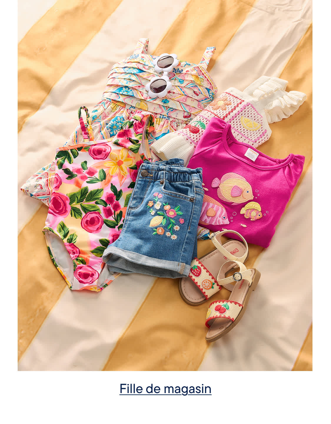 Une collection de vêtements d'été colorés pour enfants, comprenant un maillot de bain à fleurs, un short en jean brodé de fleurs, un t-shirt rose à motifs de poissons et des sandales, est disposée sur un fond de tissu rayé jaune et blanc.