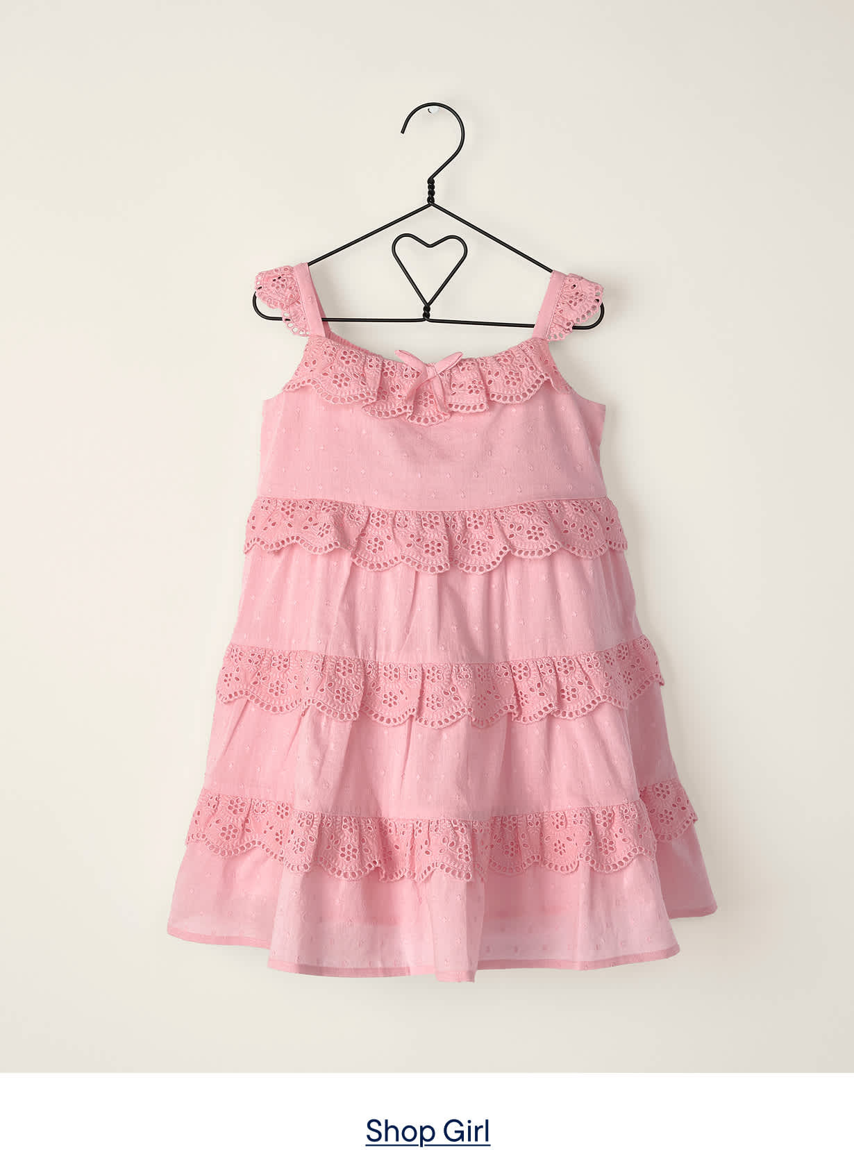 A pink, tiered, eyelet dress hangs on a heart-shaped hanger against a plain, light-colored wall.