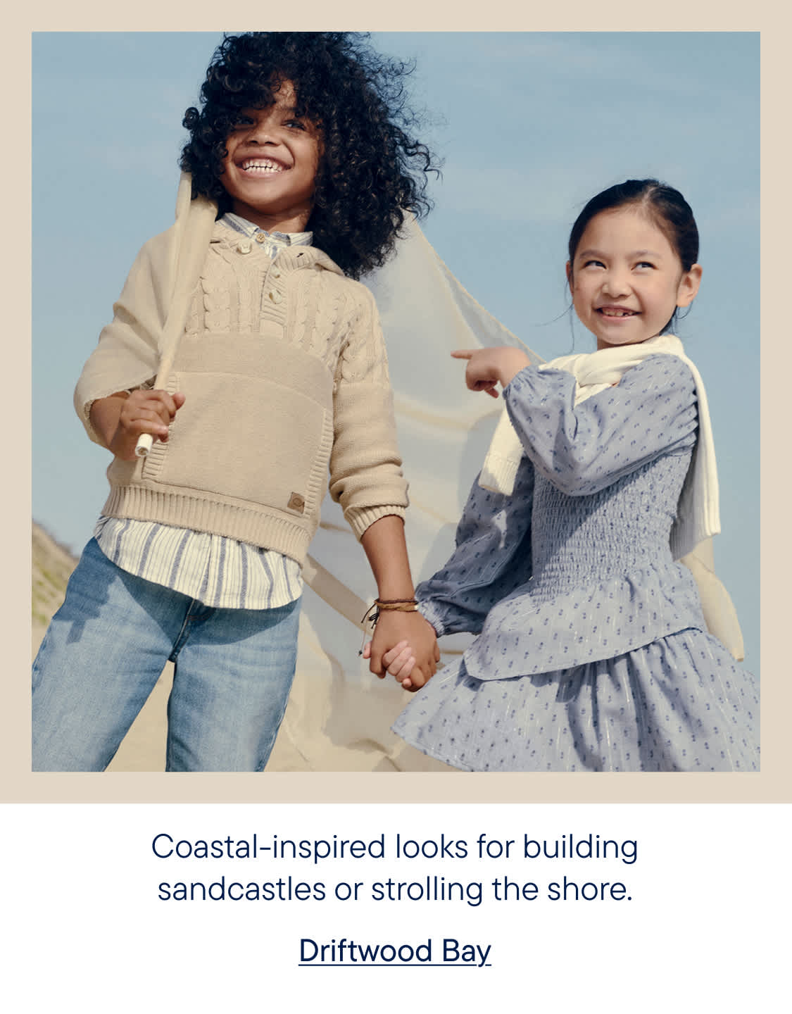 Two smiling children in casual outfits stand on a sandy beach with a sheer fabric billowing behind them, under a clear blue sky.