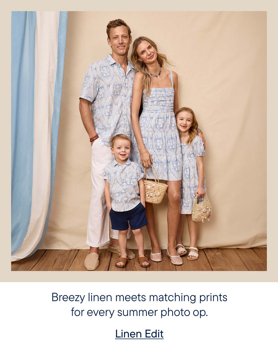 A family of four, dressed in matching blue and white patterned outfits, poses in front of a light beige backdrop with blue and white striped curtains on the left.