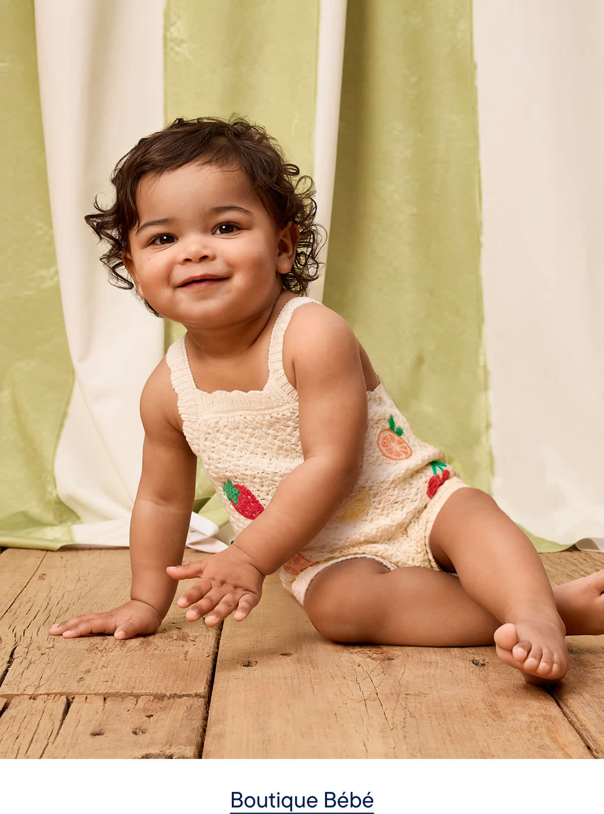 Un bébé souriant, vêtu d'une barboteuse à motifs de fruits, est assis sur un parquet devant un fond à rayures vertes et blanches.