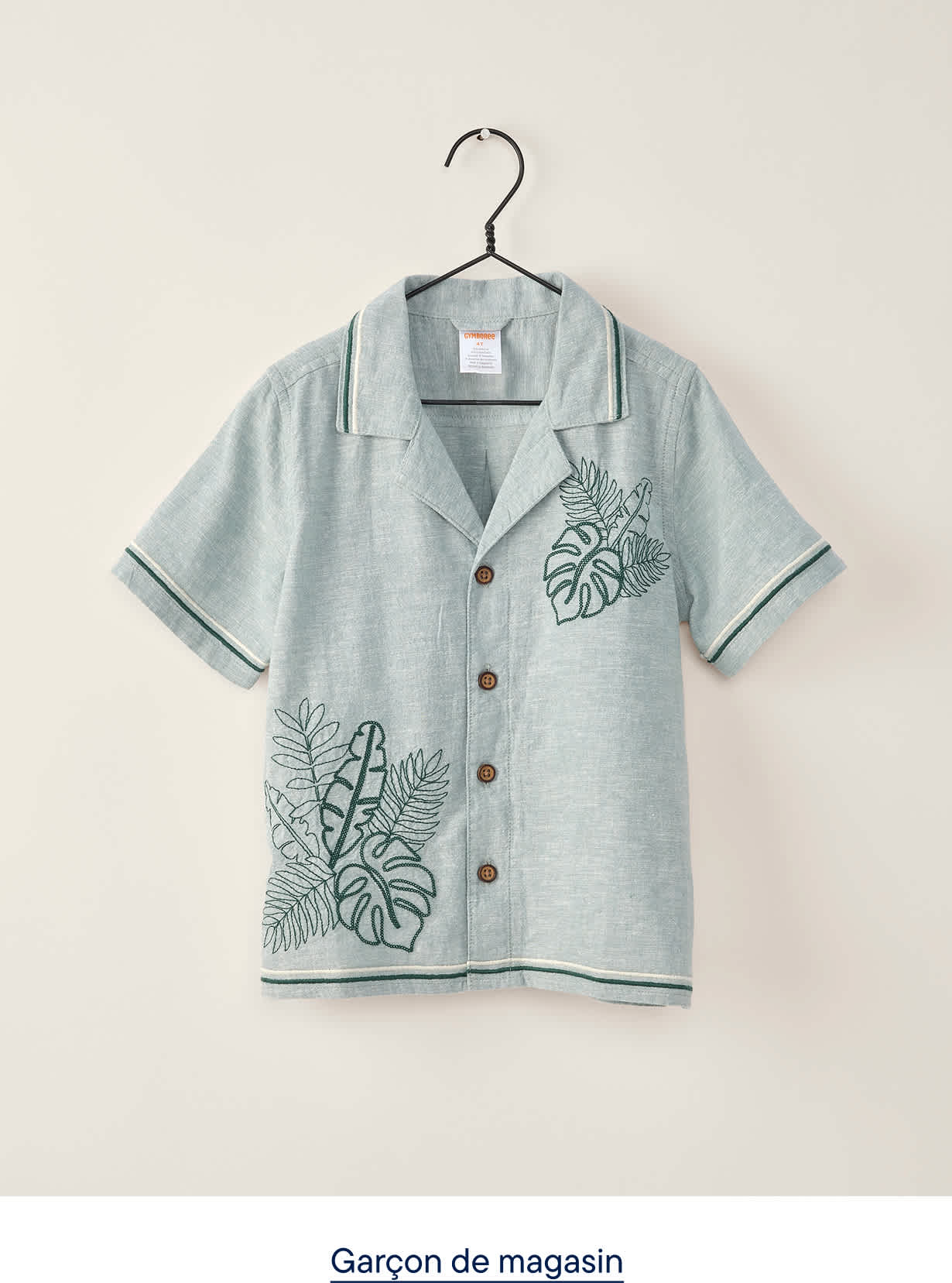 Une chemise bleu clair à manches courtes, ornée de broderies de feuilles vertes, est suspendue à un cintre noir sur un fond gris clair uni.