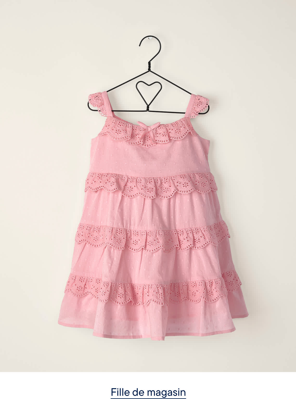 Une robe rose à volants et à œillets est suspendue à un cintre en forme de cœur contre un mur clair et uni.