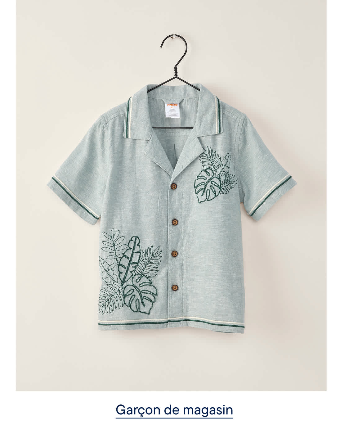 Une chemise bleu clair à manches courtes, ornée de broderies de feuilles vertes, est suspendue à un cintre sur un fond uni et clair.