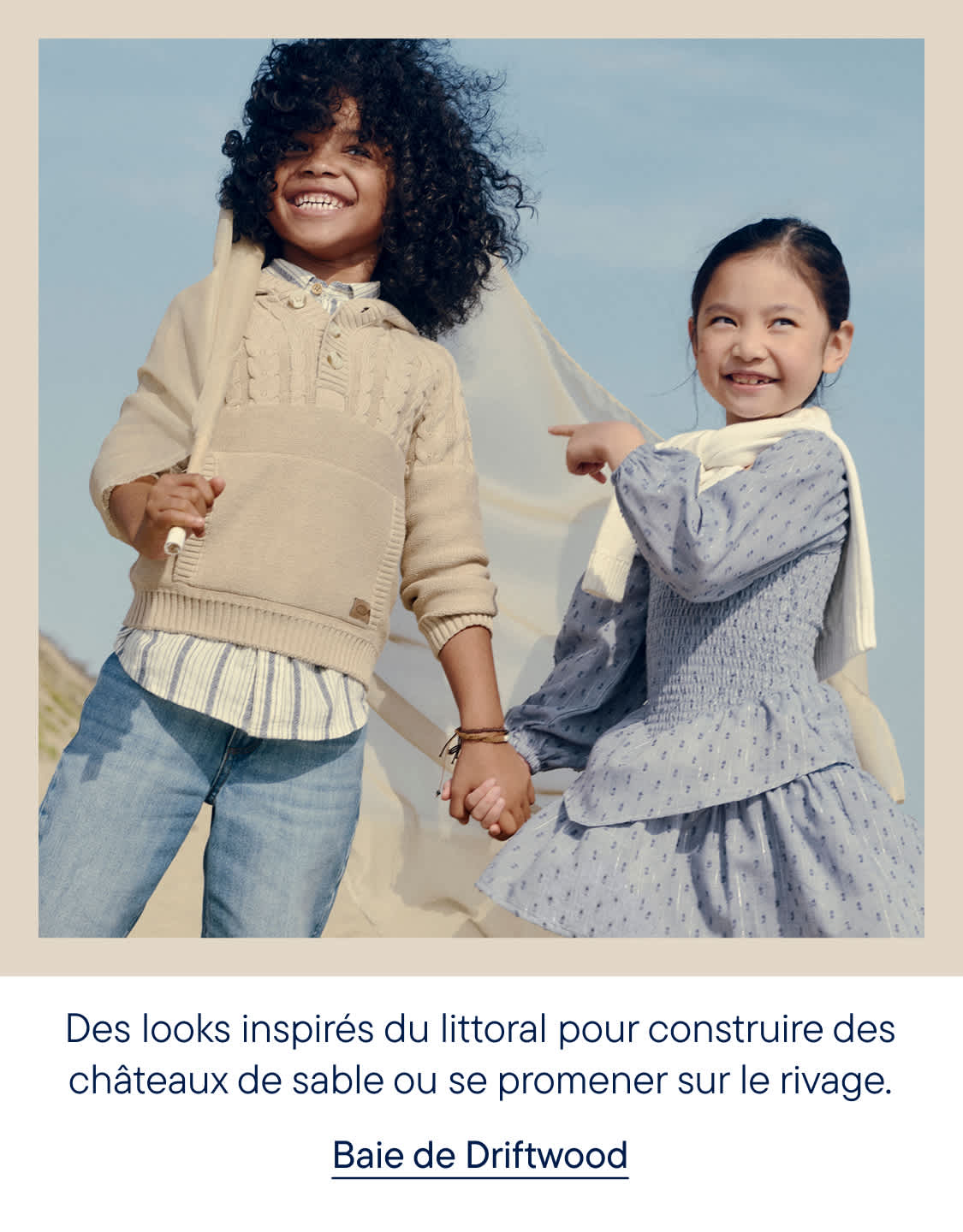 Deux enfants souriants, vêtus de tenues décontractées, se tiennent sur une plage de sable fin, un voile de tissu flottant derrière eux, sous un ciel bleu limpide.