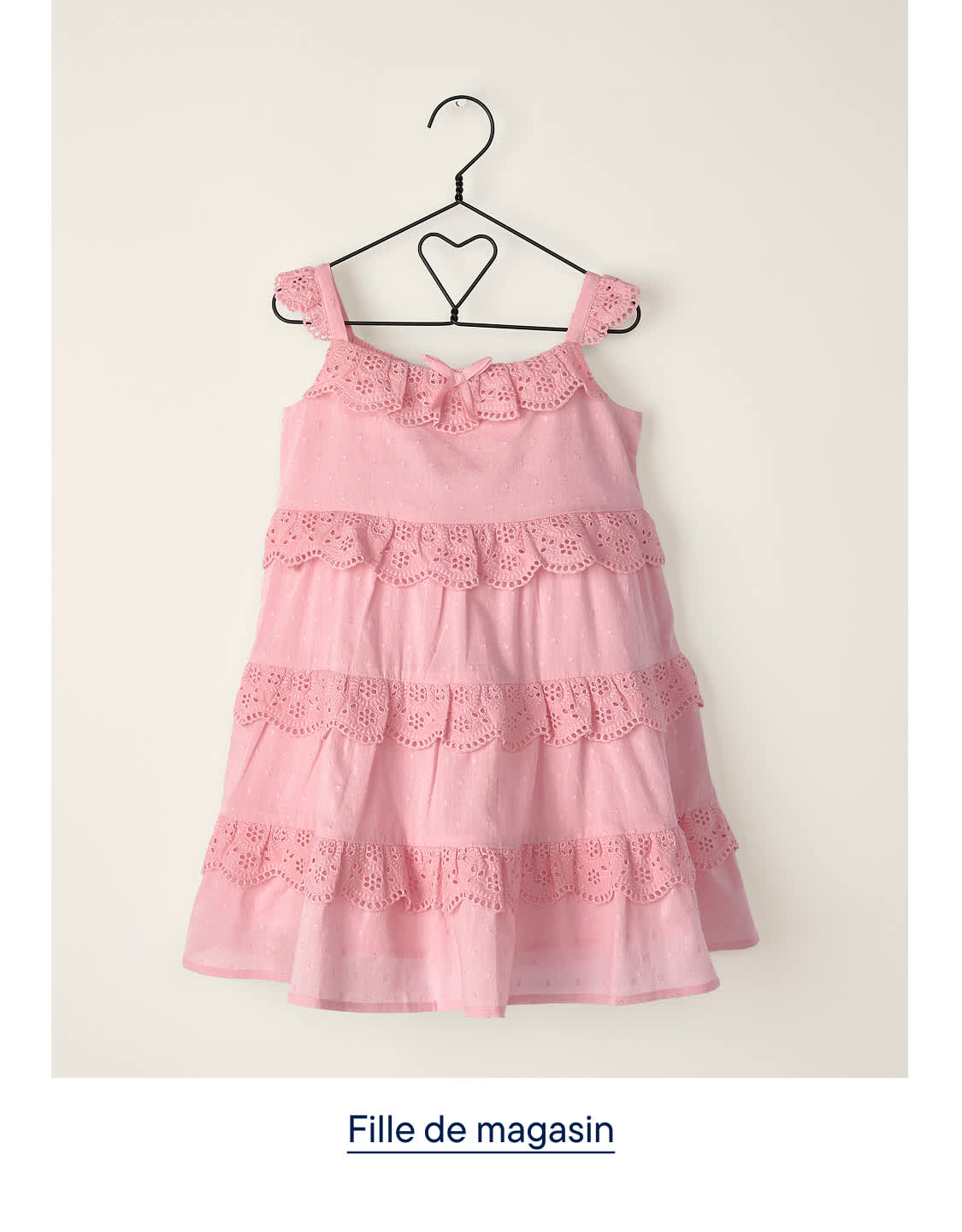 Une robe rose à volants ornée de broderies anglaises est suspendue à un cintre en forme de cœur sur un fond clair et uni.