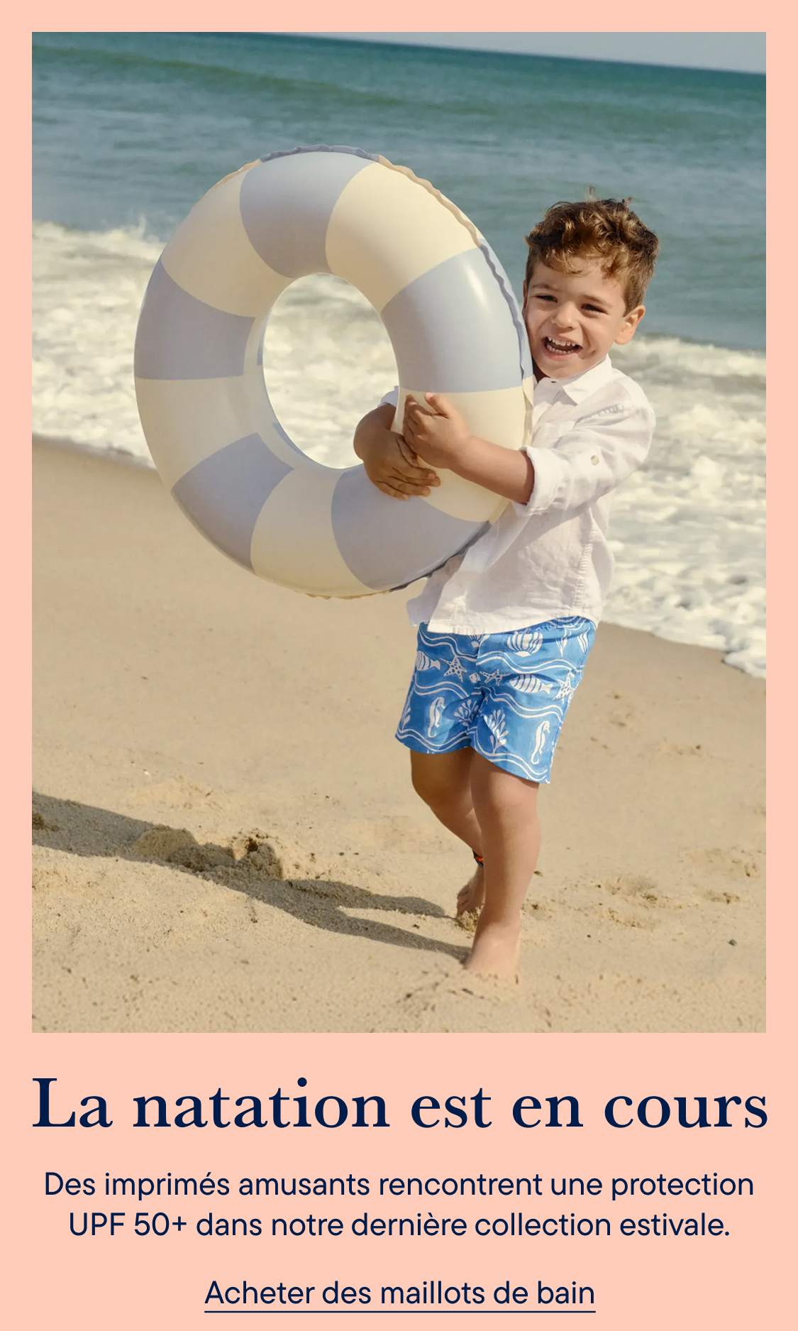 Un garçon souriant, en maillot de bain et t-shirt blanc, tient une bouée gonflable à rayures sur une plage de sable fin, avec l'océan en arrière-plan.