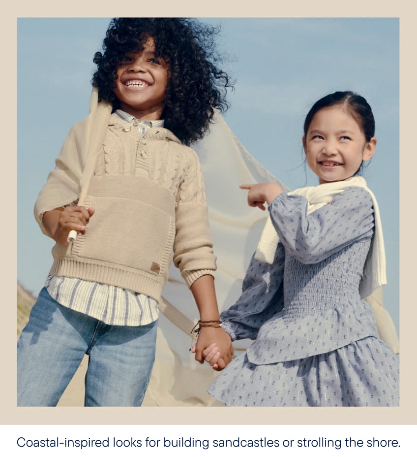 Two smiling children, a boy with curly hair and a girl with dark hair, hold hands on a sandy beach with a sheer fabric billowing behind them, under a pale blue sky.