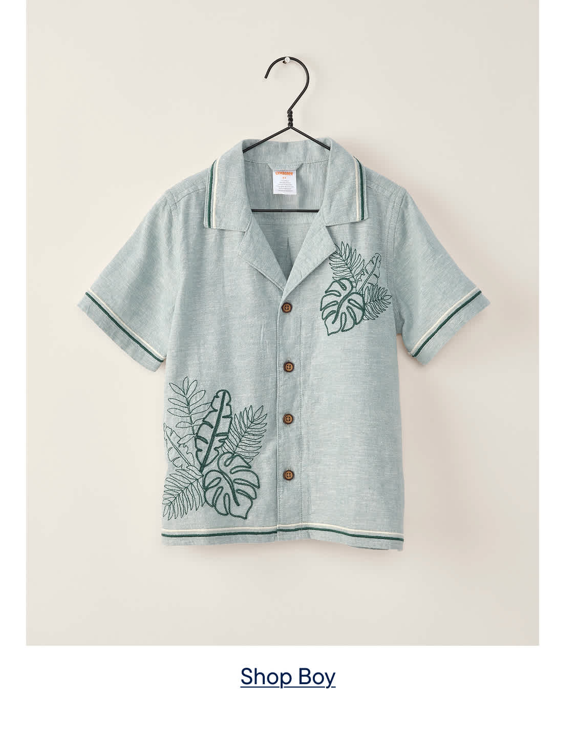 A light blue short-sleeved shirt with green leaf embroidery hangs on a hanger against a plain, light-colored background.