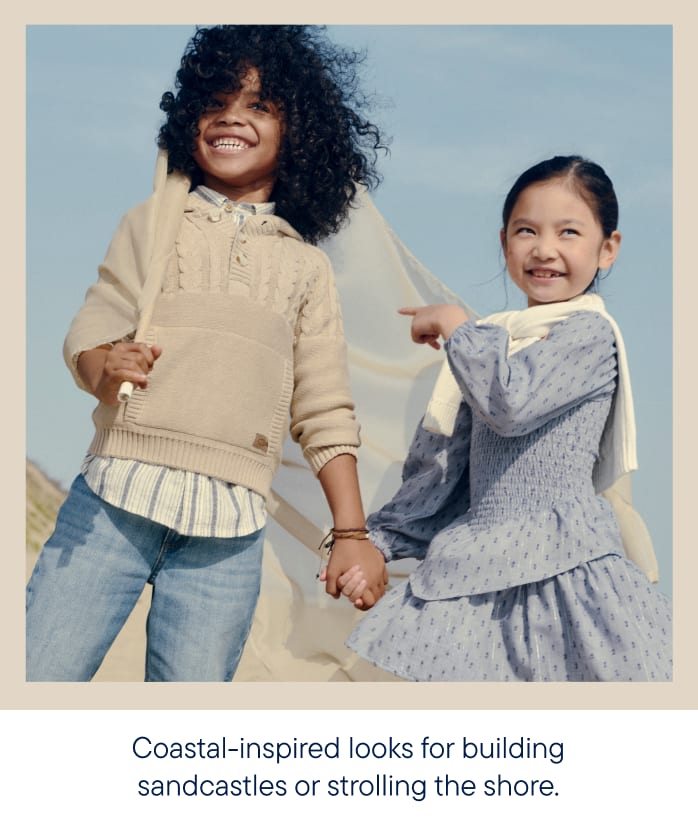 Two smiling children, a boy and a girl, stand hand-in-hand on a beach with a light blue sky in the background.