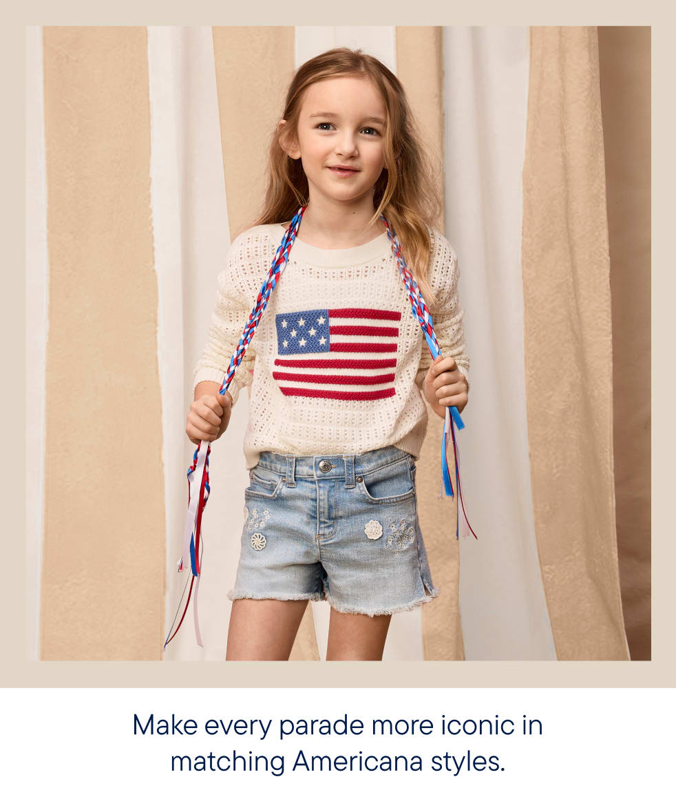 A young girl with braided hair stands in the foreground wearing a sweater with the American flag and denim shorts, against a background of beige and white striped fabric.