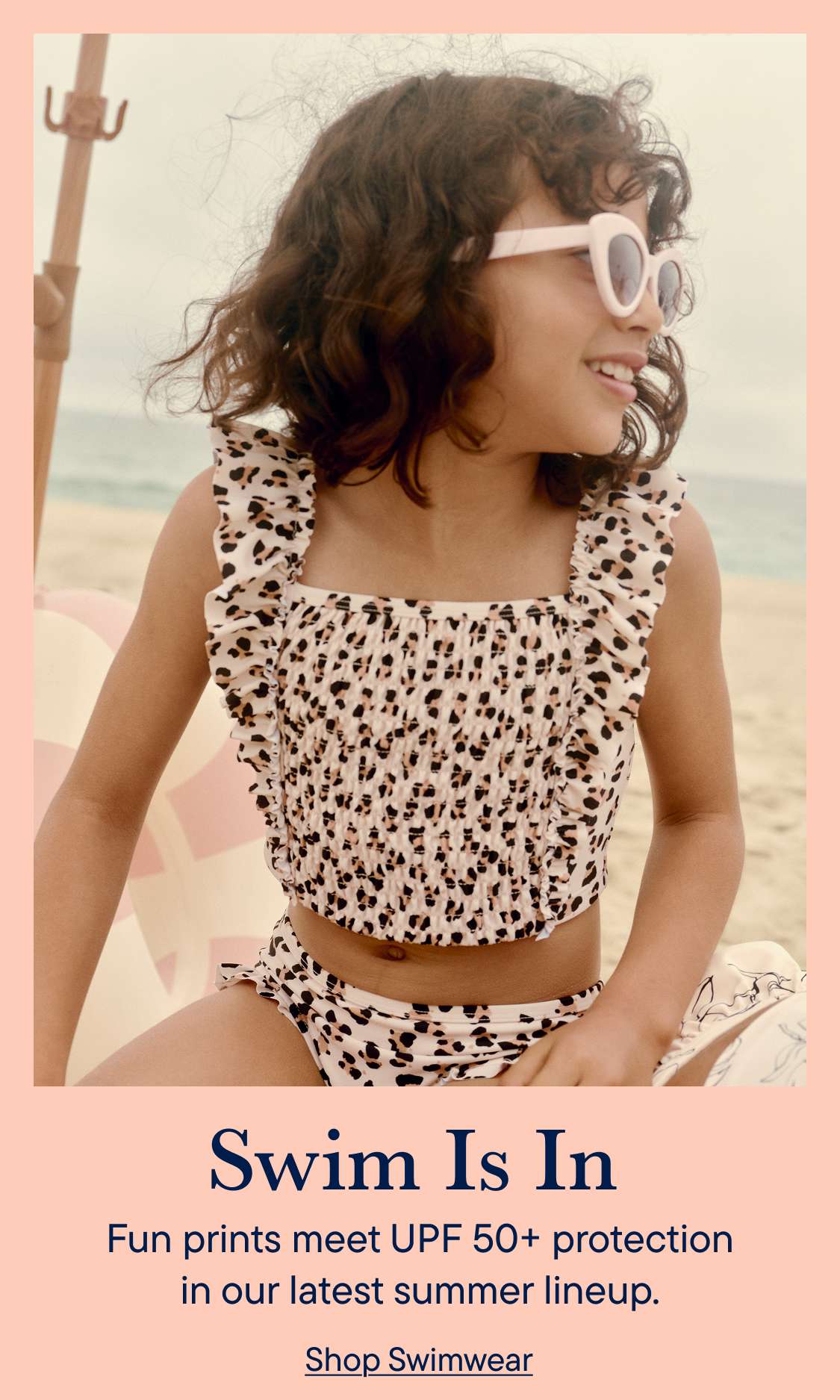A young girl in a leopard-print bikini smiles while sitting on a beach with the ocean in the background.