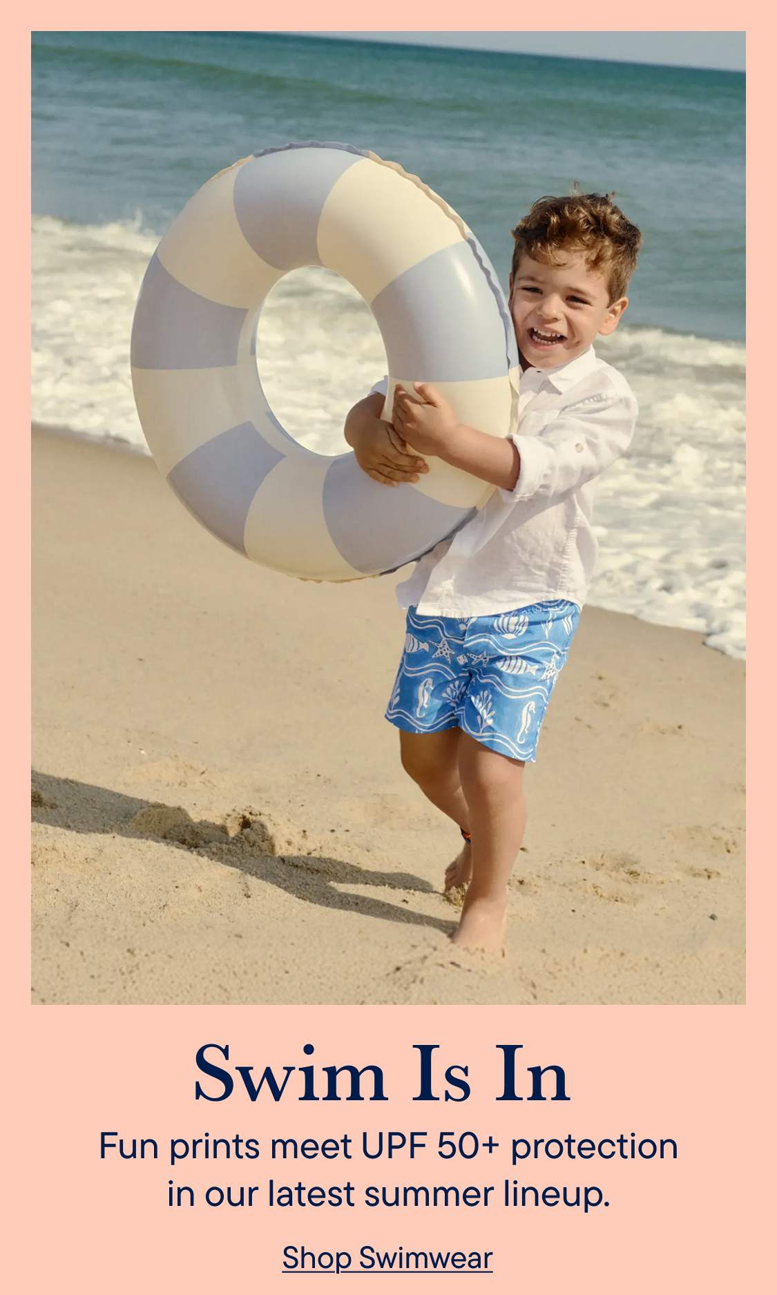A smiling boy in swim trunks and a white shirt holds a striped inflatable ring on a sandy beach with the ocean in the background.