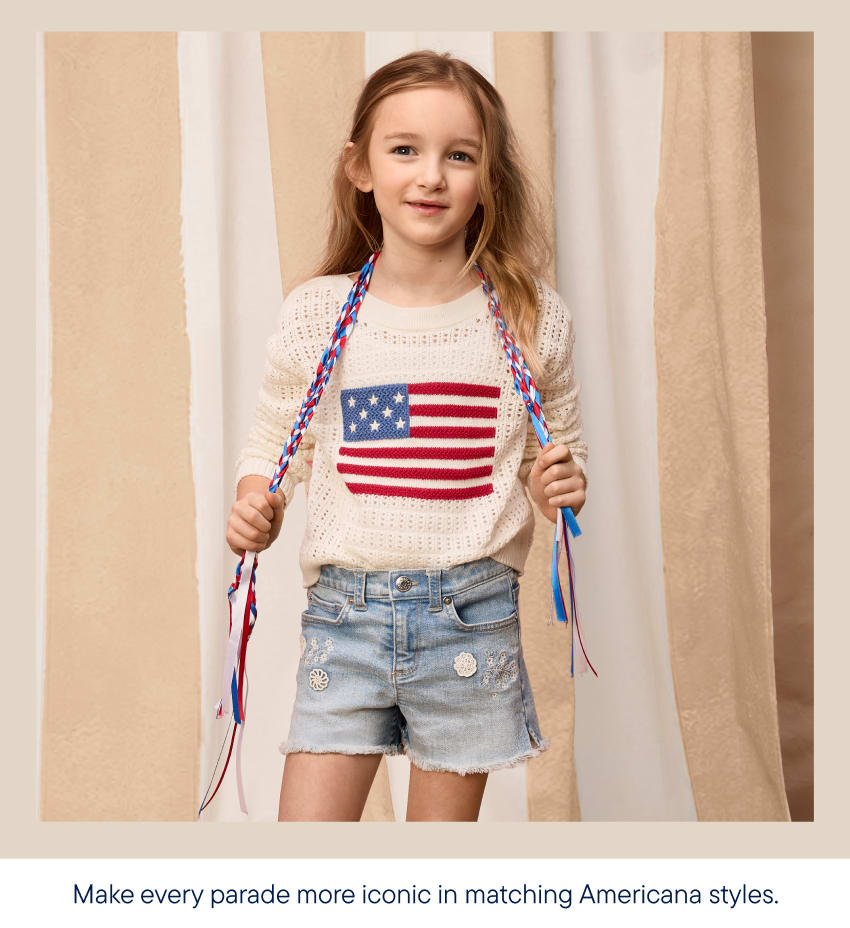 A young girl with braided hair adorned with red, white, and blue ribbons stands in the foreground, wearing a cream-colored sweater with an American flag design and denim shorts, against a backdrop of beige and white striped curtains.