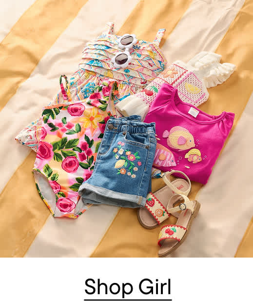 A collection of colorful children's summer clothes, including a swimsuit, shorts, and a t-shirt, are laid out on a striped yellow and white blanket.