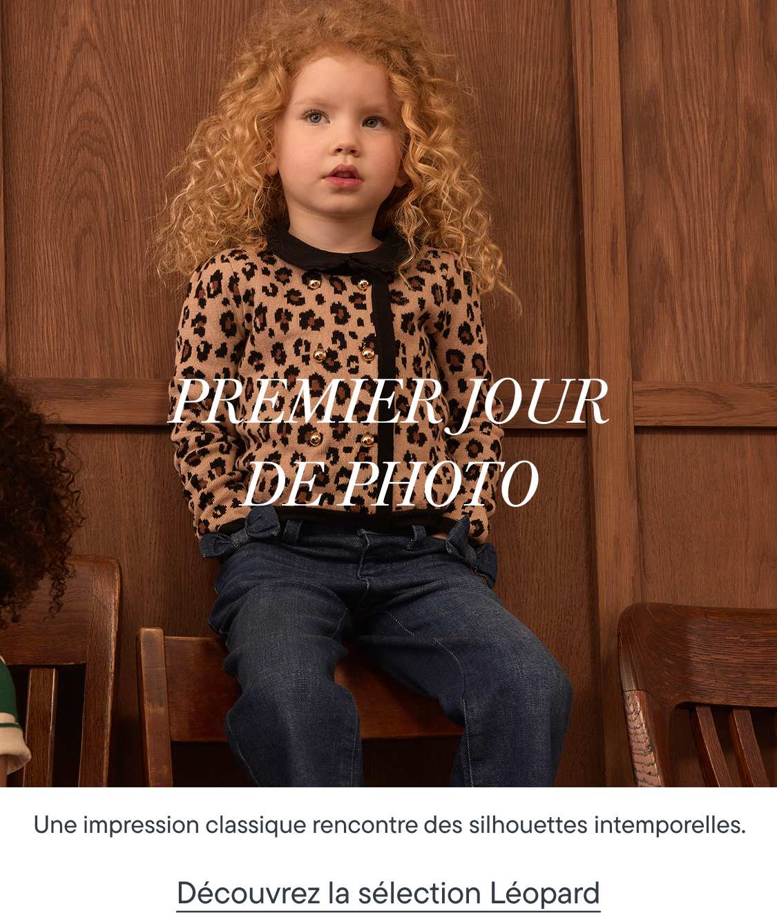 Une jeune fille aux cheveux blonds bouclés, vêtue d'un cardigan à imprimé léopard et d'un jean, est assise sur une chaise en bois devant un mur lambrissé.