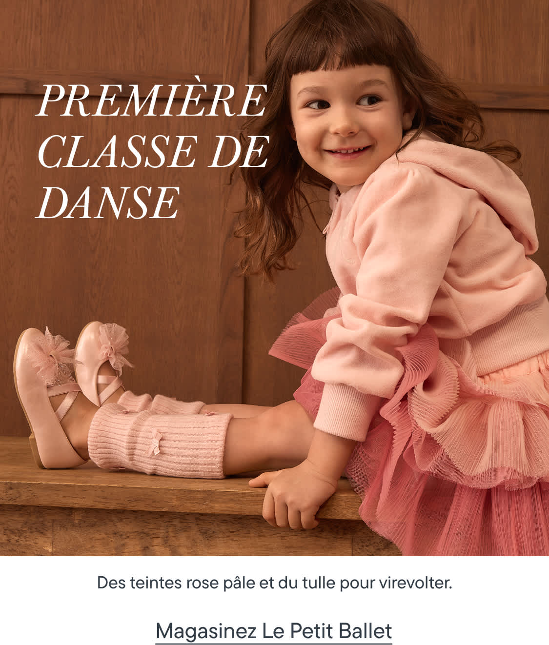 Une jeune fille en tutu rose et chaussons de ballet est assise sur un banc en bois devant un fond lambrissé.