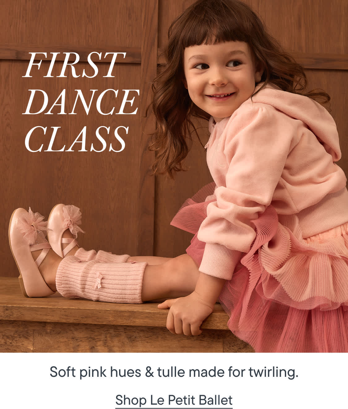 Una niña pequeña con un tutú rosa y zapatillas de ballet está sentada en un banco de madera frente a un fondo revestido de paneles de madera.