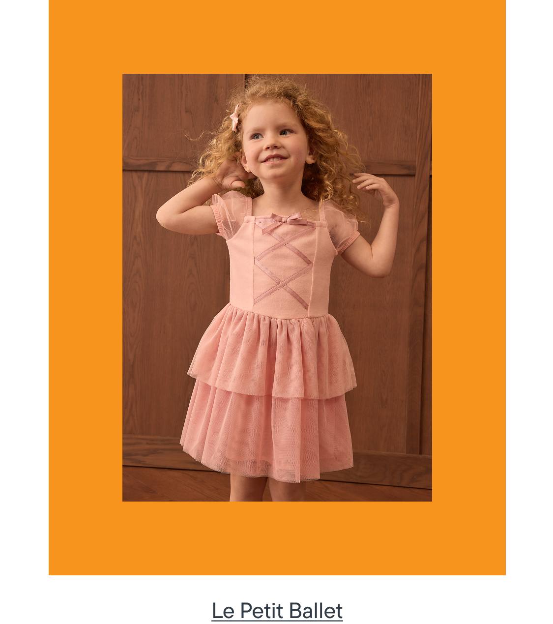 A young girl in a pink ballet dress stands in front of a wooden door, framed by an orange border.