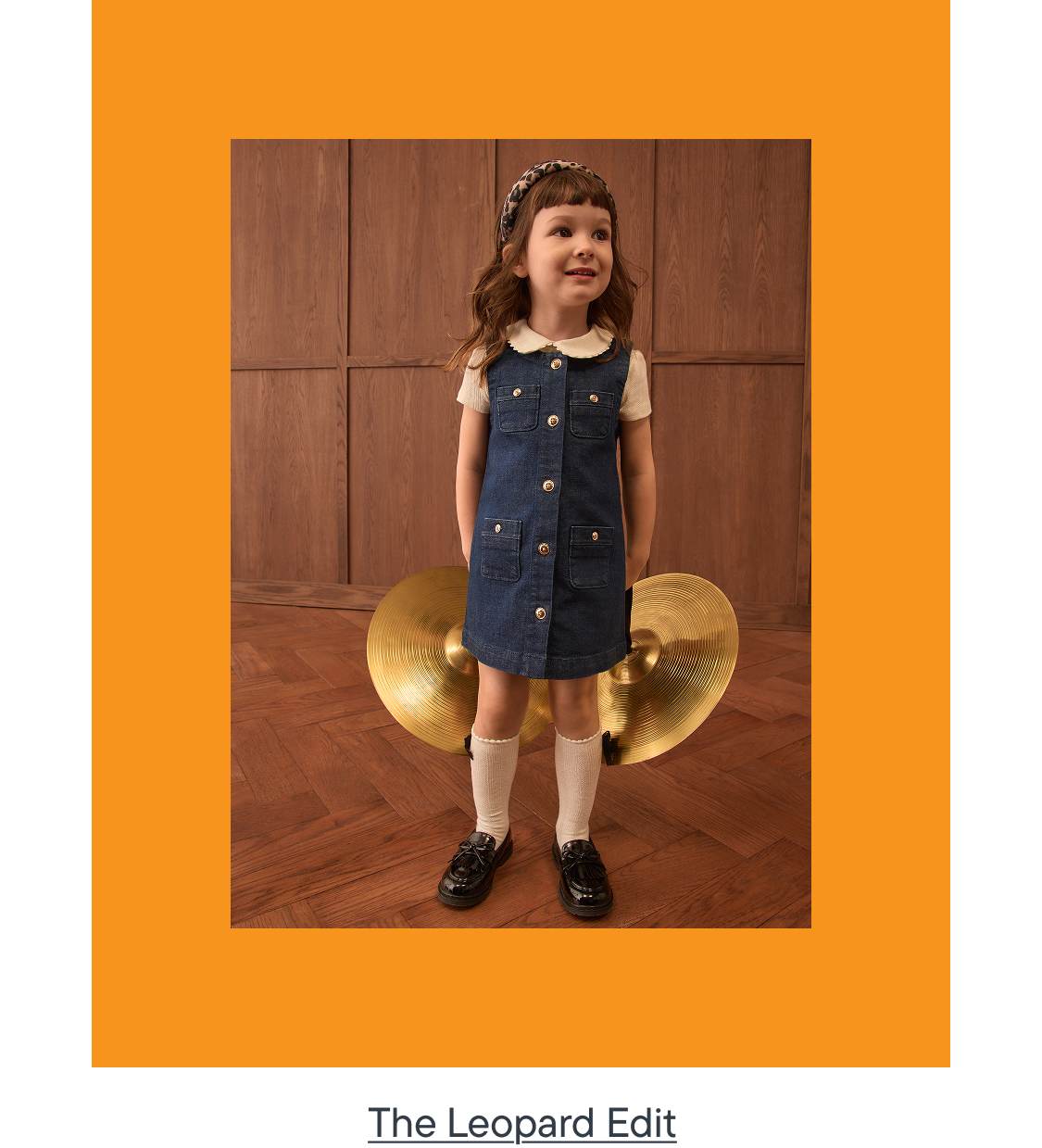 A young girl in a denim dress stands in front of two large cymbals, framed by an orange border.