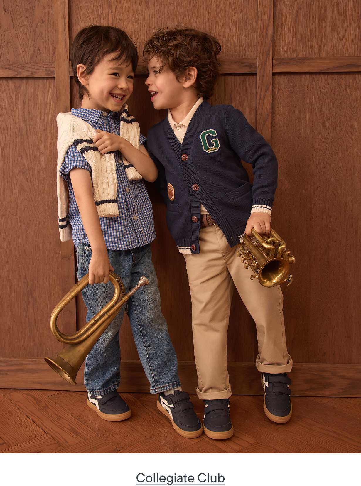 Two young boys in collegiate-style clothing, one holding a trumpet, stand against a warm, wood-paneled background.