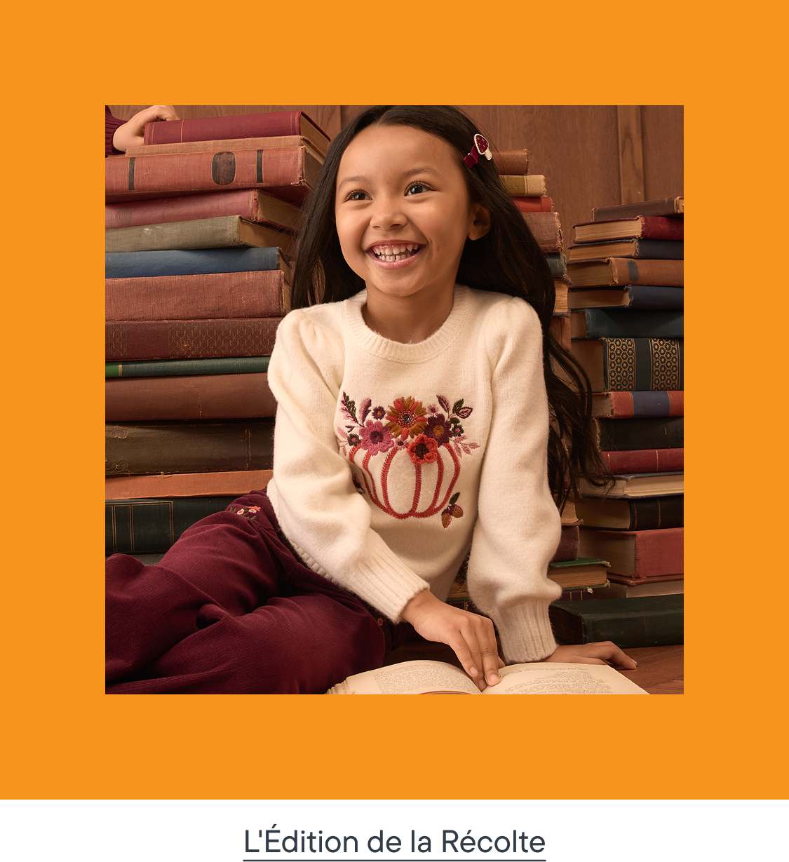 Une jeune fille souriante, vêtue d'un pull brodé de citrouilles, est assise par terre, un livre ouvert à la main, entourée de piles de vieux livres.