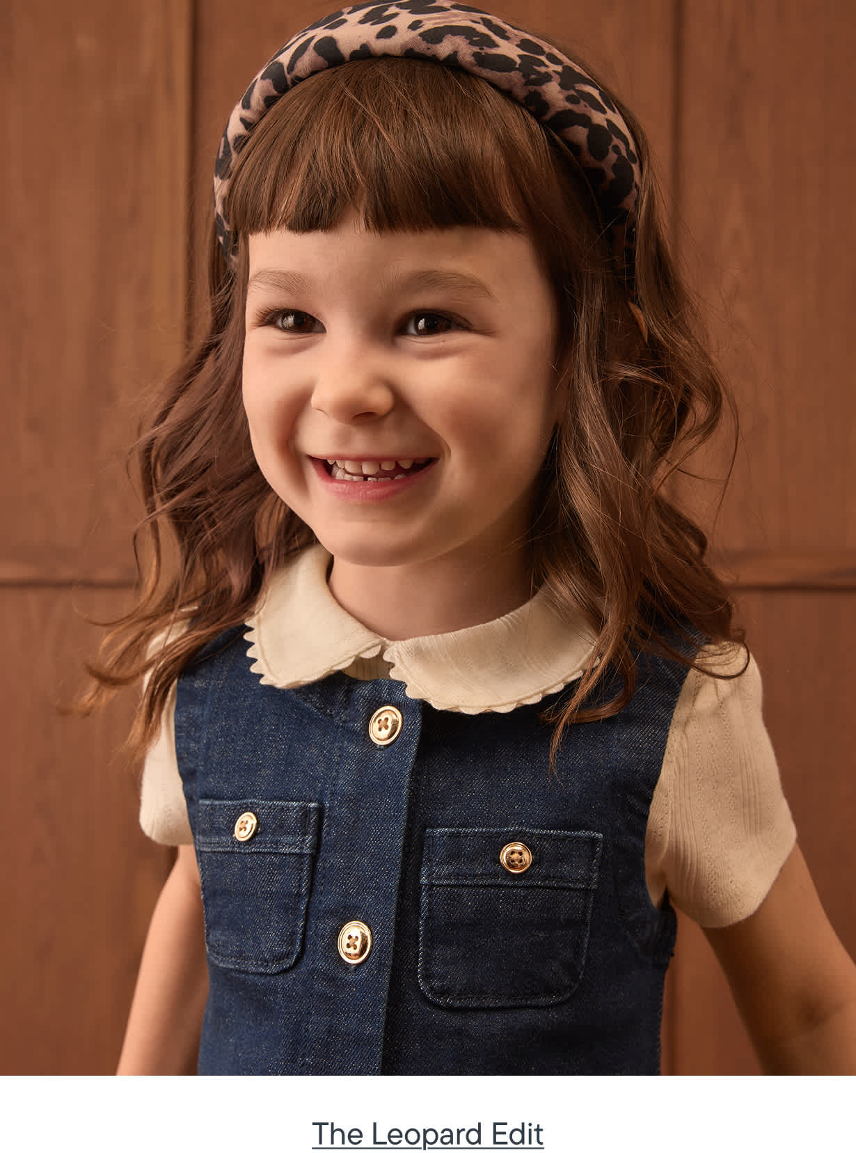A smiling young girl with a leopard print headband and denim dress stands against a warm, wood-paneled background.