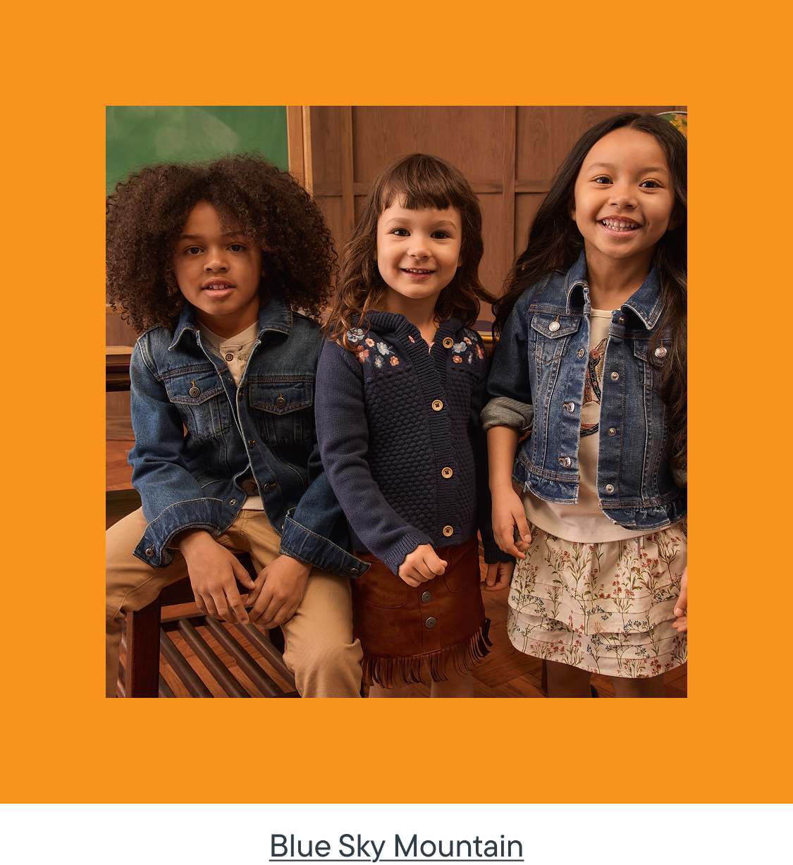 Three smiling children in casual outfits pose in front of a wooden background.