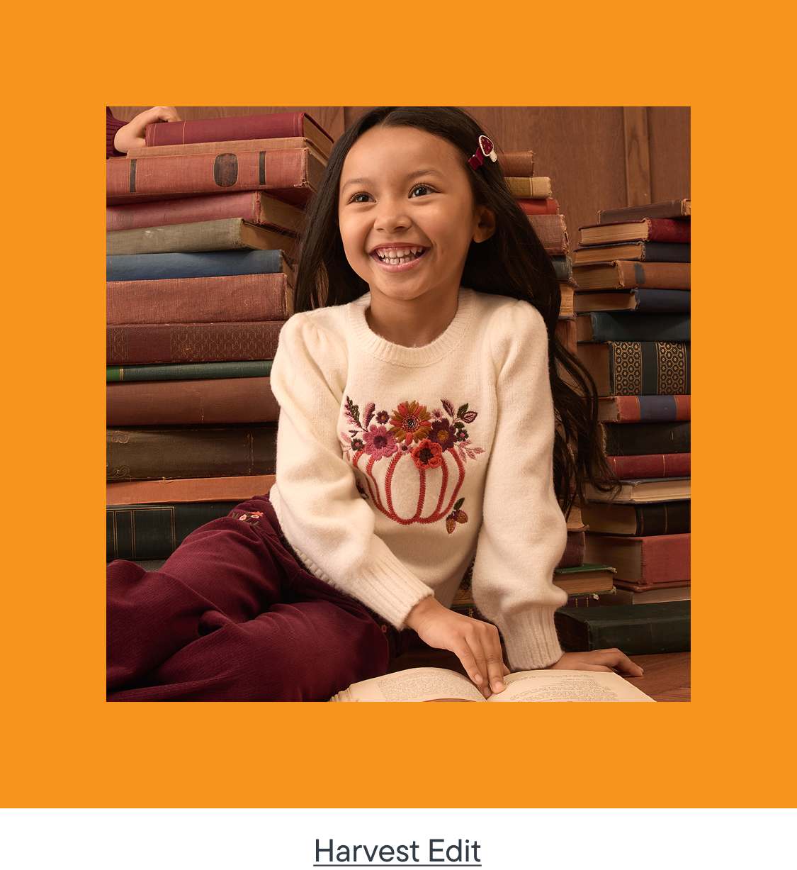 A smiling young girl in a pumpkin-embroidered sweater sits on the floor with an open book, while a tall stack of old books forms the background.